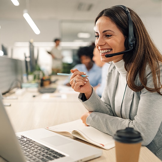 Smiling caucasian business woman with headset working on a laptop in a call center on the line. Happy female working in a busy customer service department and taking notes while talking with a client.